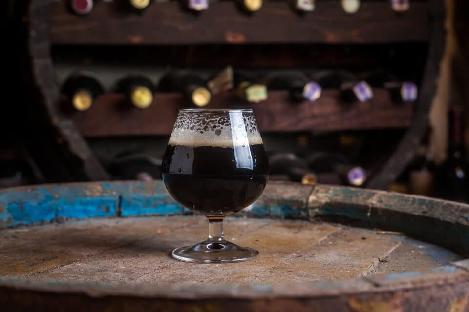 Snifter glass full of dark ale standing on a wooden barrel in a cellar.