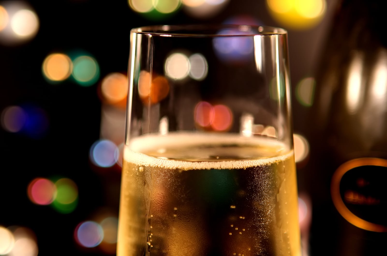 Closeup of a glass of bubbling champagne, against multi-colored lights. Bottle in background.